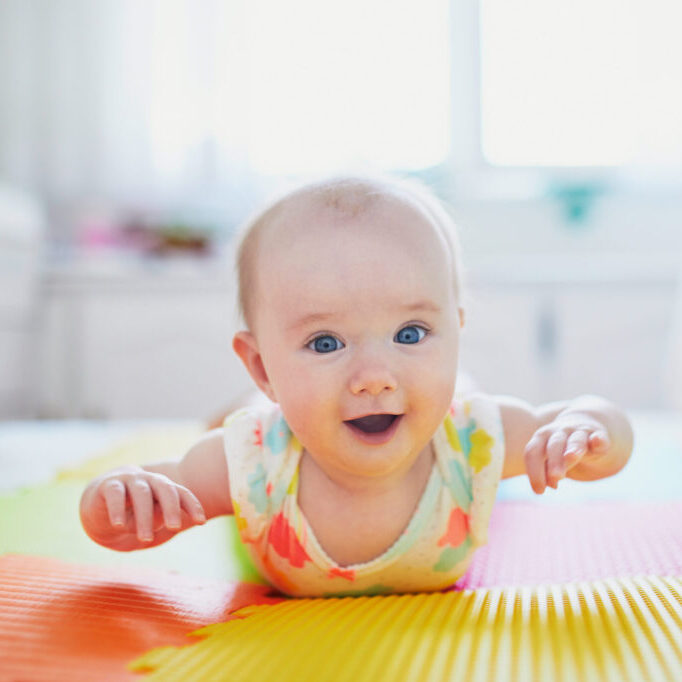 Happy smiling baby girl lying on colorful play mat on the floor and trying to crawl