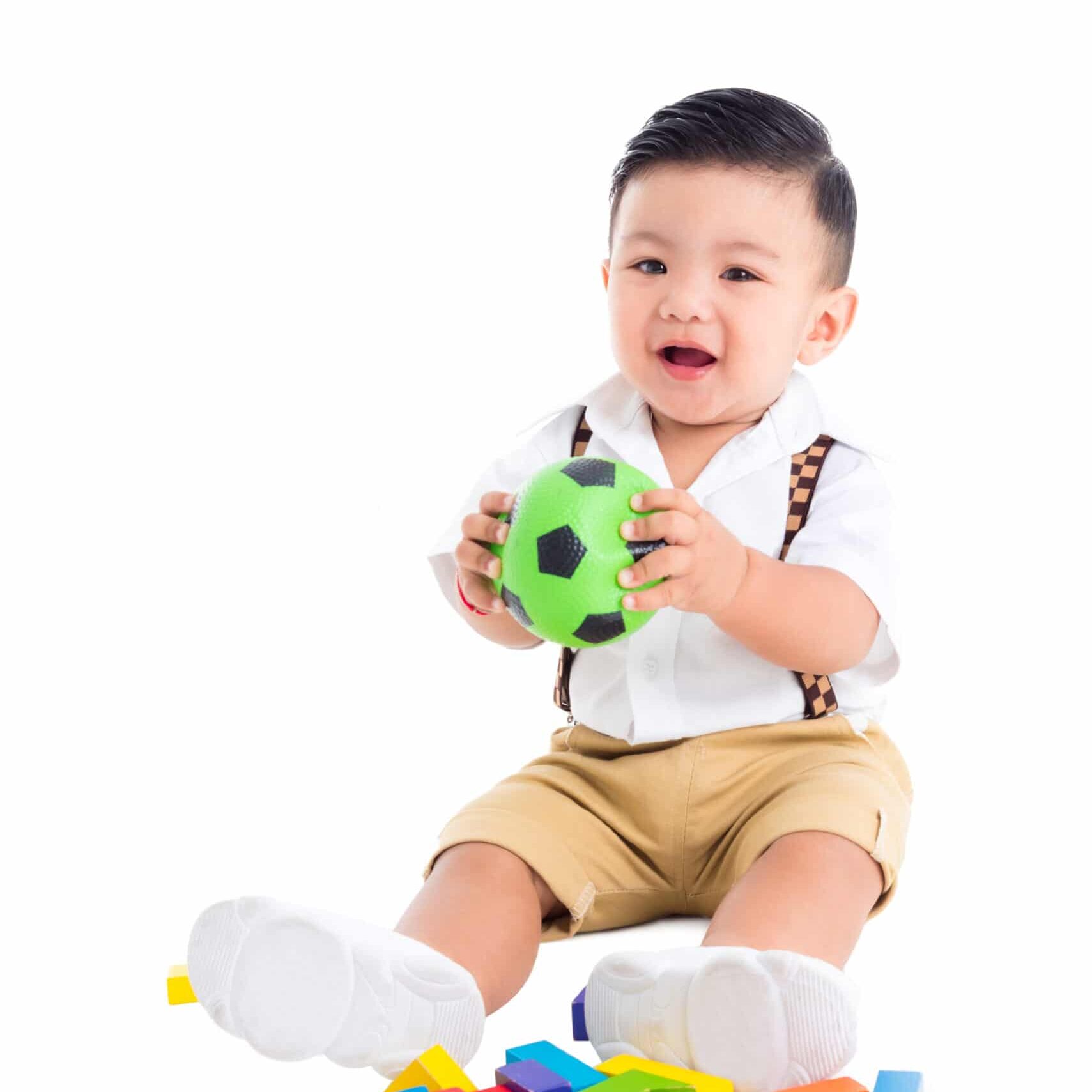 Little asian male toddler sitting on the floor and holding ball and smiles over white background